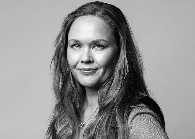 Kvinna med långt hår, klädd i väst och långärmad skjorta, tittar in i kameran mot en enfärgad bakgrund.