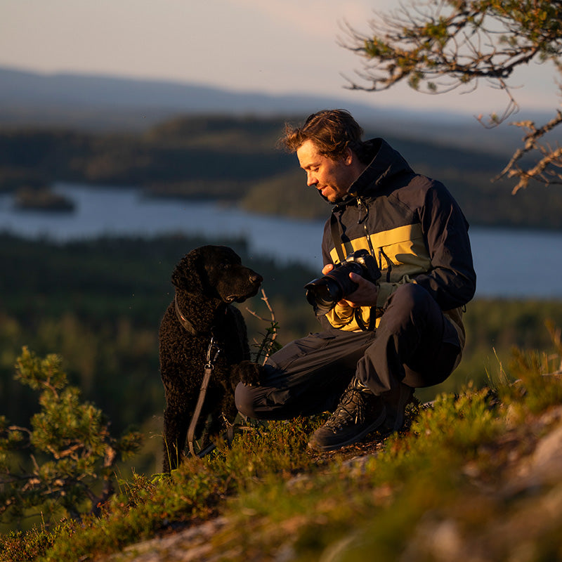 En man i friluftskläder står på knä i gräset med en kamera i handen och tittar på en svart hund med en sjö och träd i bakgrunden.