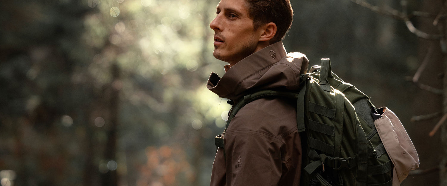 En man i brun jacka och grön ryggsäck står utomhus i en solbelyst skog och tittar åt sidan.