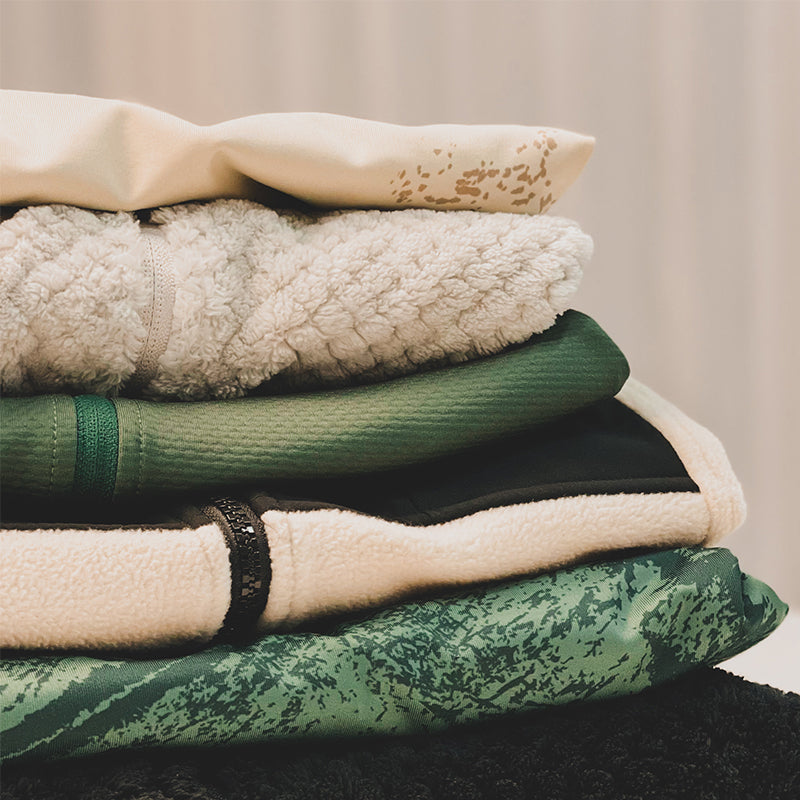 Närbild på en hög med vikta kläder med olika texturer och nyanser av grönt, vitt och svart.