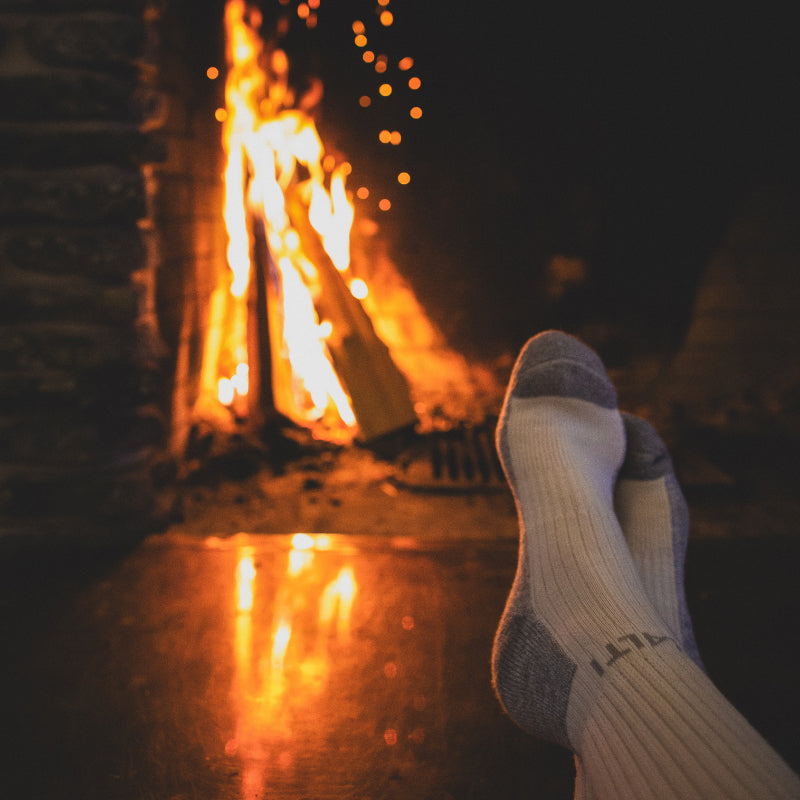 En person i vita strumpor slappnar av med fötterna upp framför en tänd öppen spis med glödande lågor och gnistor.