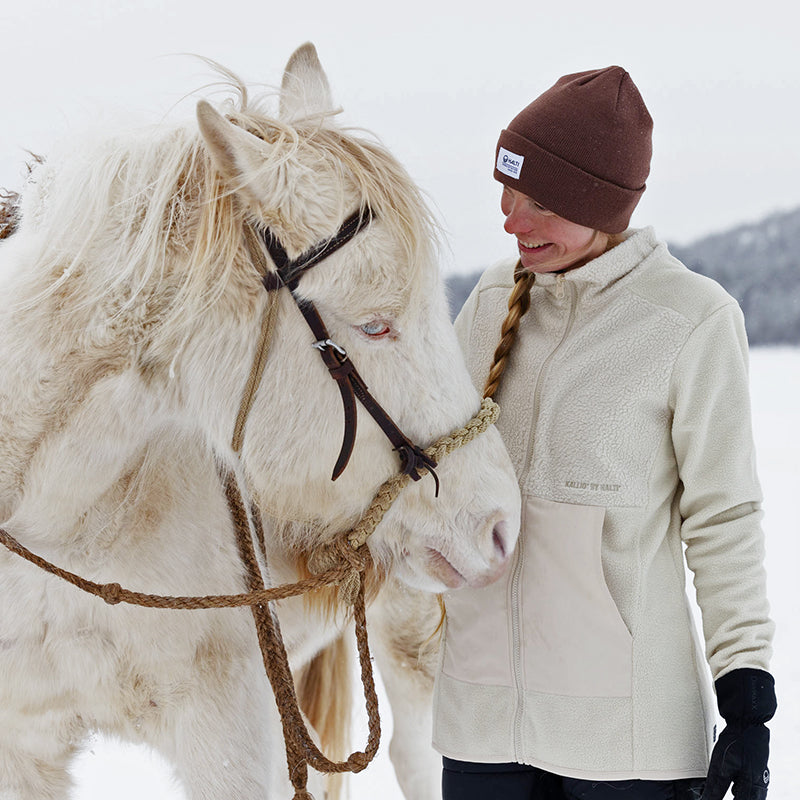 En kvinna i brun mössa och gräddfärgad jacka står bredvid en ljus häst utomhus i snöigt väder.