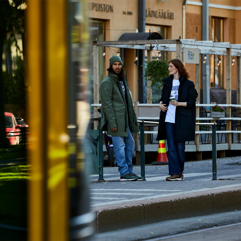 Två personer står på en trottoar i en stad nära ett räcke, och i förgrunden passerar ett gult fordon.