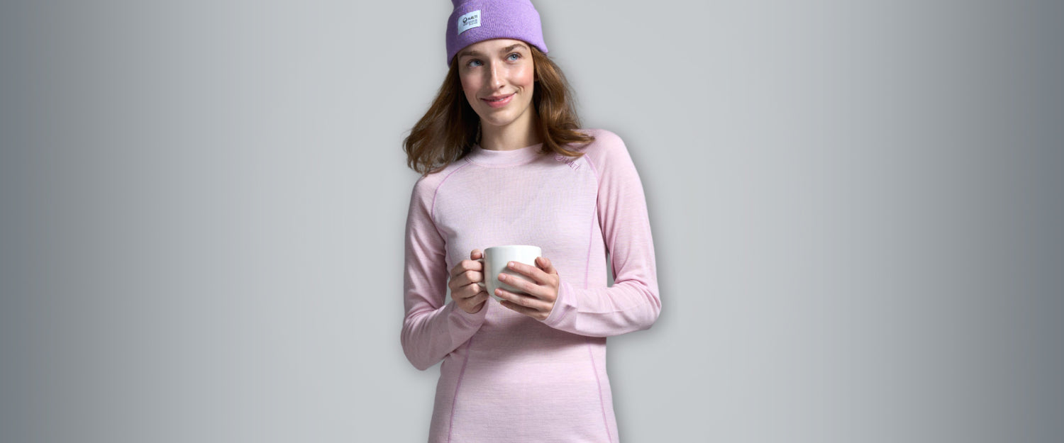 En person i rosa långärmad tröja och lila mössa håller i en vit mugg mot en grå bakgrund.