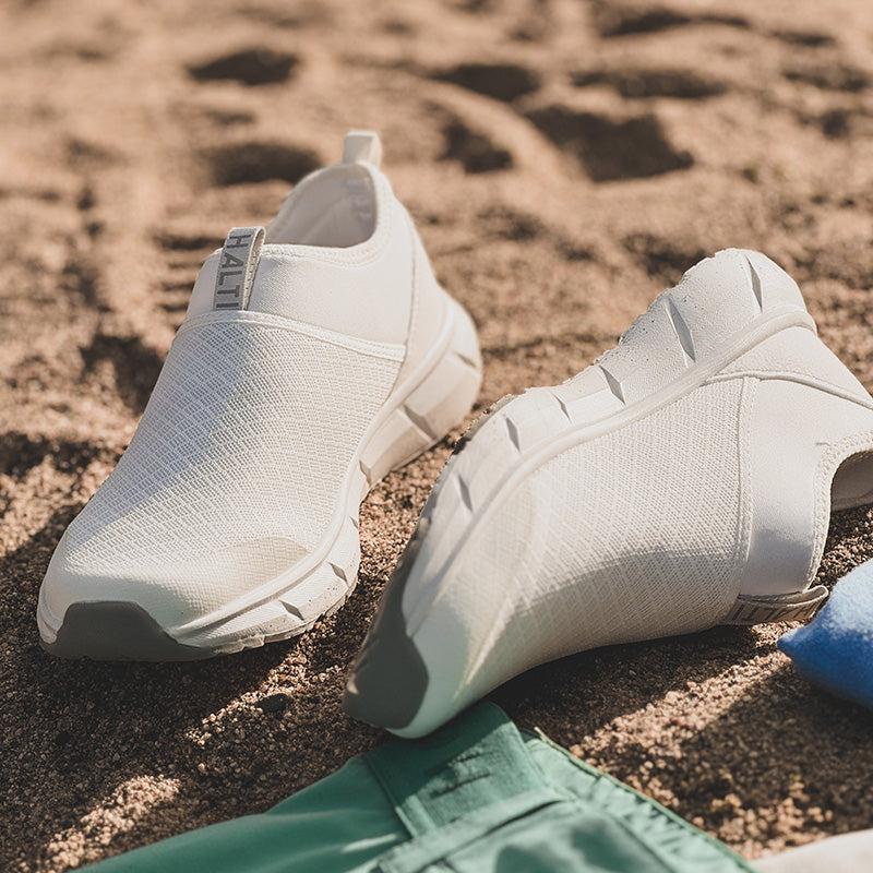 Ett par vita slip-on-atletskor på sandig mark bredvid gröna shorts och ett blått föremål.