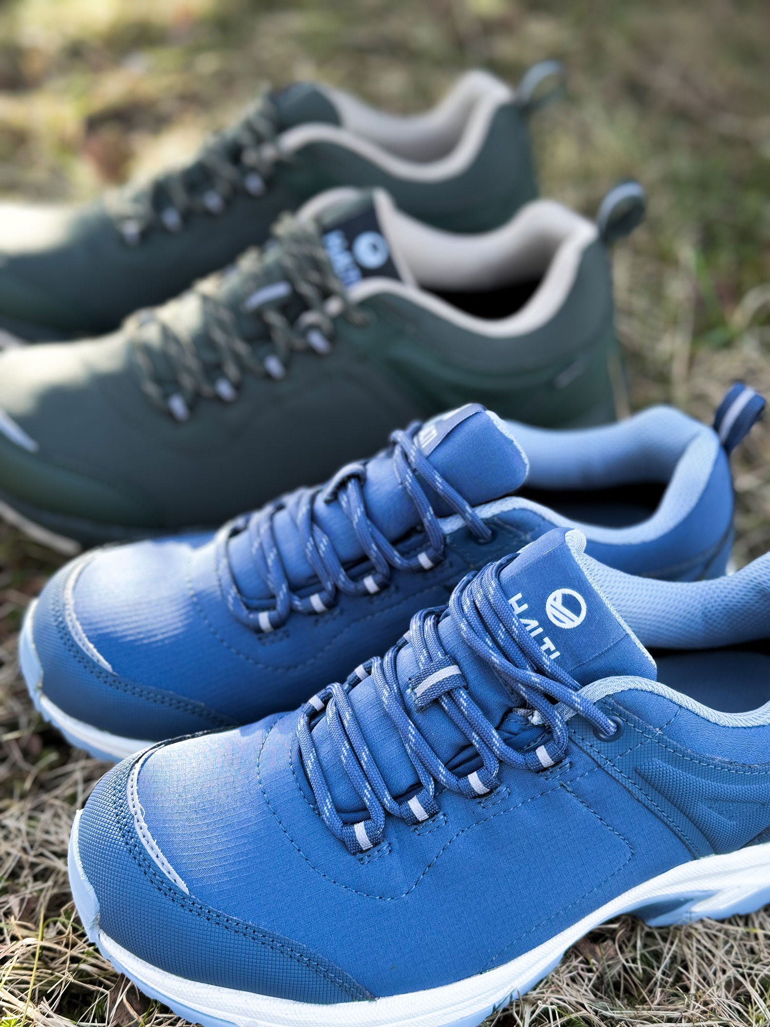 Två par Halti Outdoorskor DX Low Dam, blå respektive gröna, på gräs i naturligt ljus.