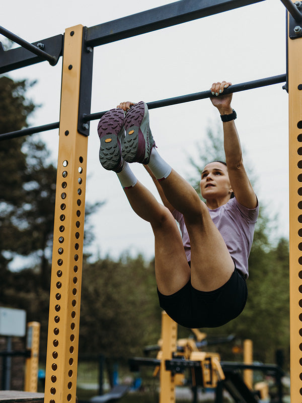 En person utför ett hängande benlyft på en pull-up-stång utomhus, klädd i lila skjorta, svarta shorts och sneakers.
