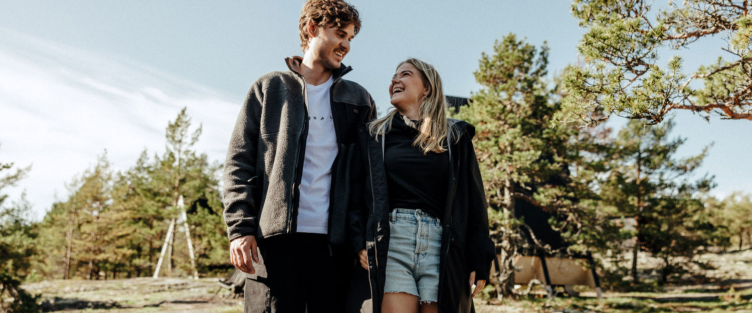 En man och en kvinna går utomhus i fritidskläder och ler mot varandra, med träd och ett tält i bakgrunden.