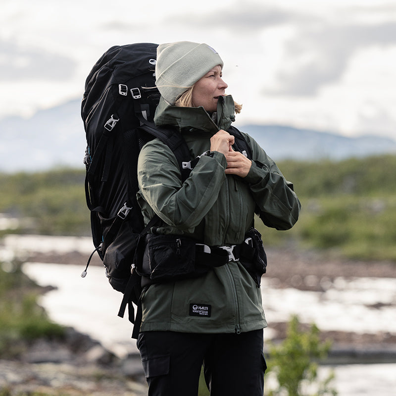 En person i friluftskläder med stor ryggsäck står vid en flod och justerar sin jacka, med berg i bakgrunden.