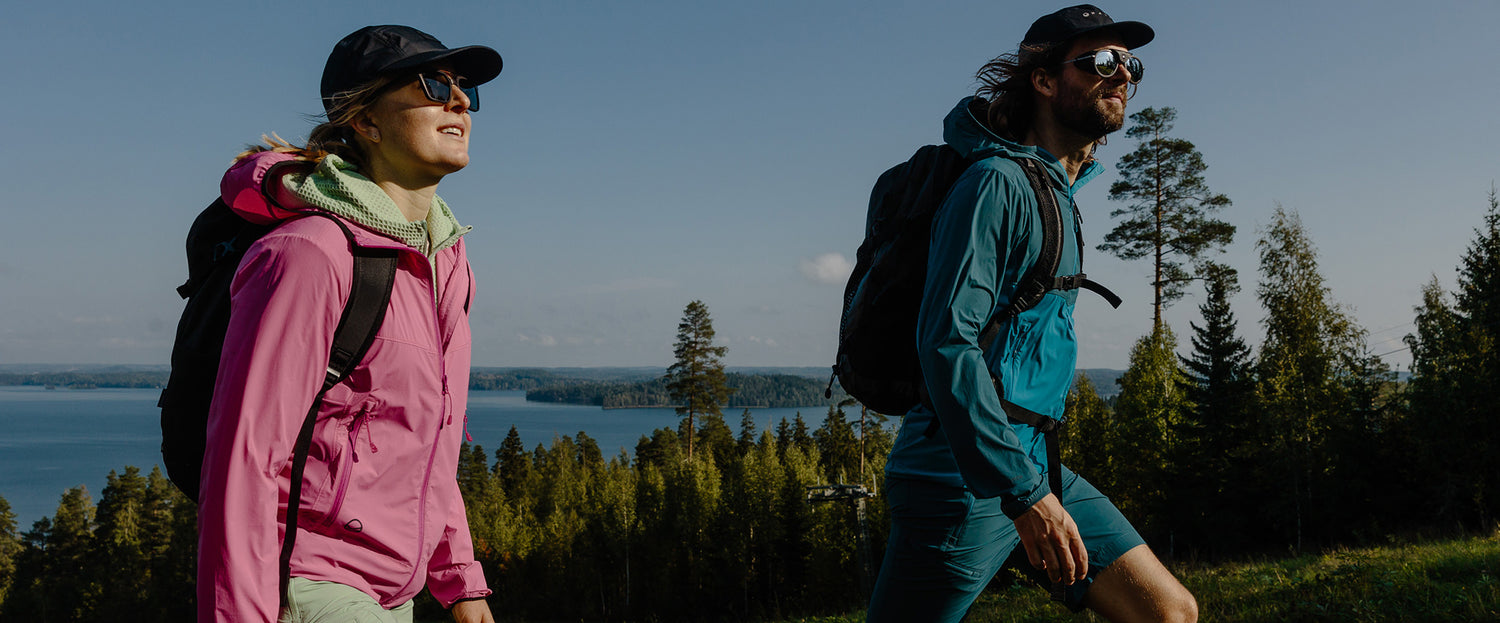 Två personer i jackor, hattar och ryggsäckar vandrar uppför en backe med träd och en sjö i bakgrunden under en klar himmel.