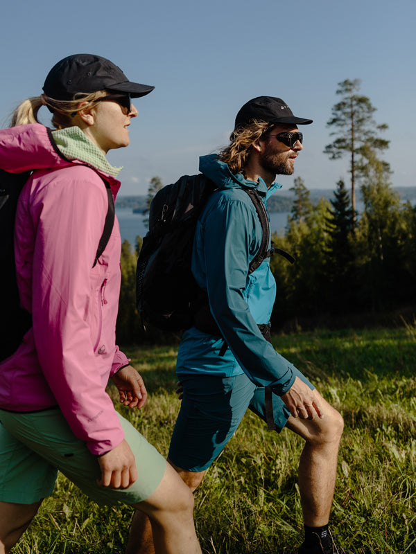 Två personer i friluftskläder och hattar vandrar uppför en gräsbevuxen stig med träd och vatten i bakgrunden.