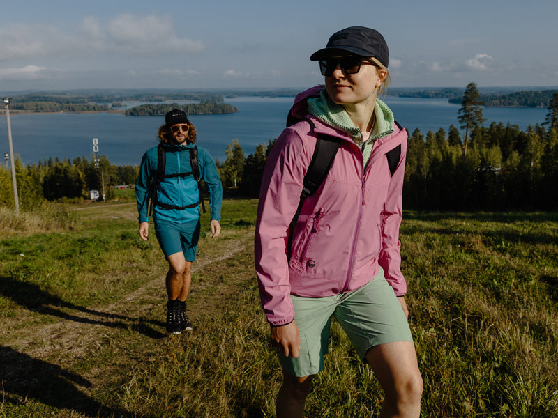 Två personer iförda friluftskläder och ryggsäckar vandrar på en gräsbevuxen stig med en sjö och skog i bakgrunden.