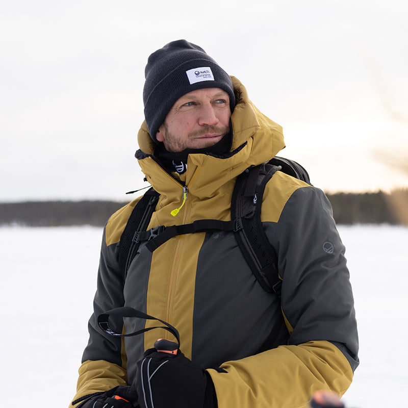 En man i vinterjacka, mössa och handskar står utomhus i ett snöigt landskap och håller i skidstavar.