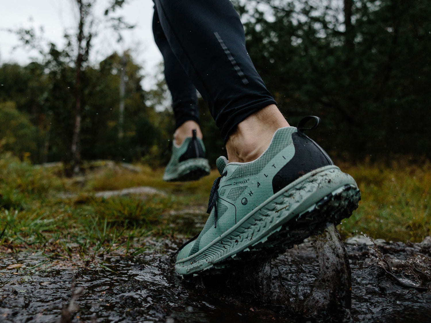 Person i gröna trailrunningskor går genom lera på en skogsstig, våt mark och träd syns i bakgrunden.