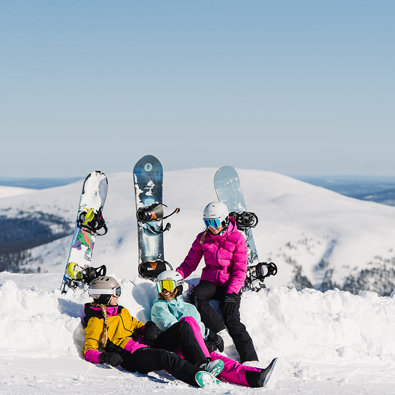Tre personer i vinterkläder med snowboards sitter och står på ett snöigt berg med klar himmel och avlägsna toppar.