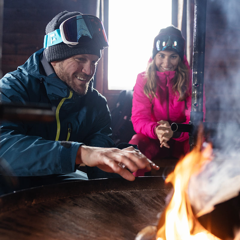 En man i skidkläder värmer sina händer vid en brasa inomhus medan en kvinna i rosa vinterkläder sitter bakom honom.