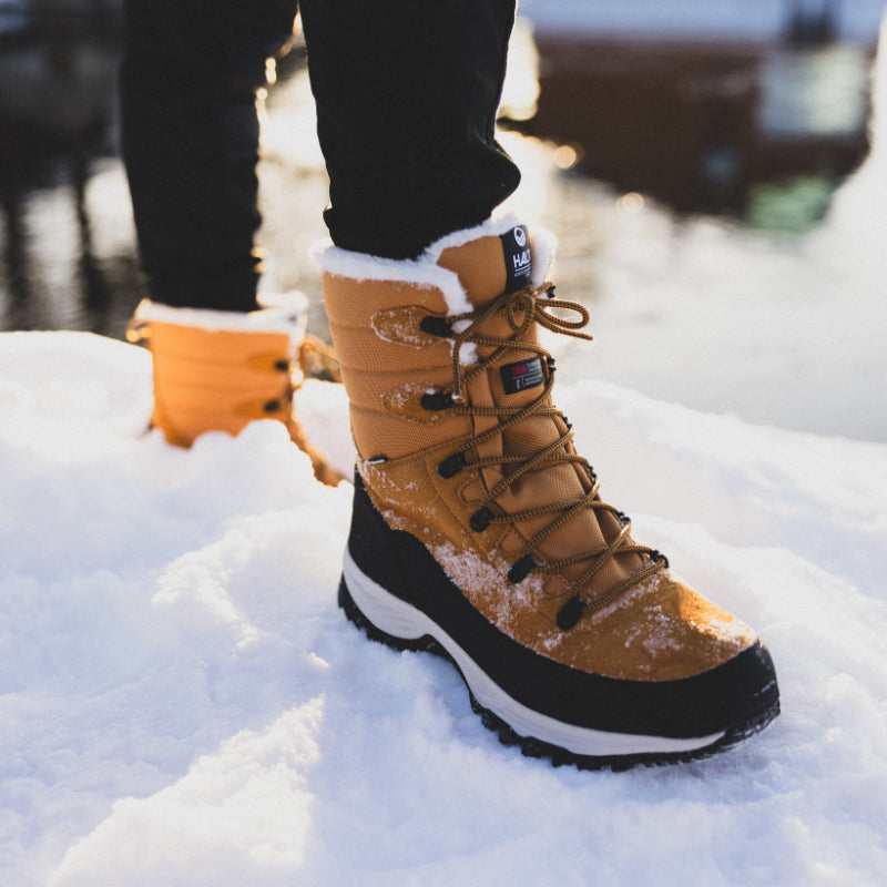 En person i solbruna och svarta vinterkängor står på snö nära ett vattendrag.