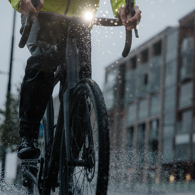 En cyklist cyklar i regn på en stadsgata, vatten stänker upp från den våta trottoaren.