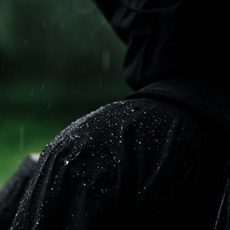 Närbild på en person som bär en svart regnjacka med vattendroppar på axeln, utomhus i regnigt väder.