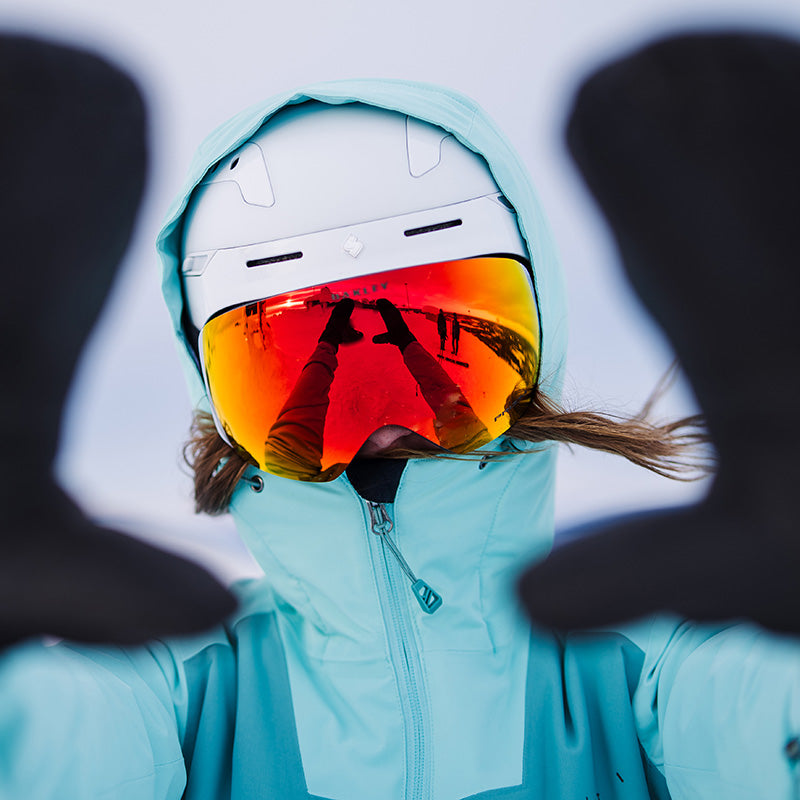 Person i blågrön jacka, hjälm och orange skidglasögon håller upp handskbeklädda händer, med reflektion synlig i glasögonen.