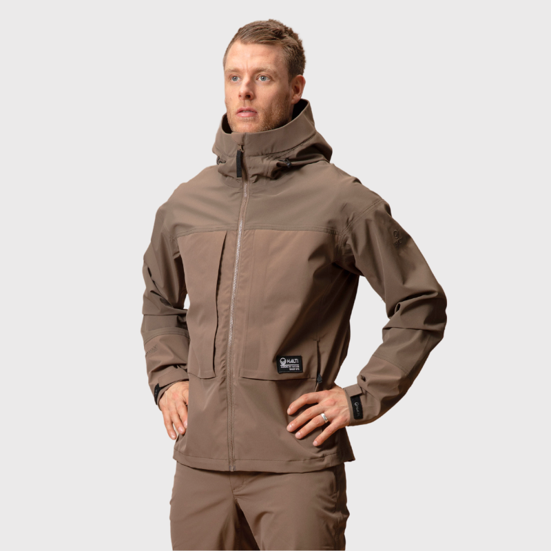 En man i brun Halti Hiker II DX Skaljacka Herr och byxor står med händerna på höfterna mot en ljus bakgrund.