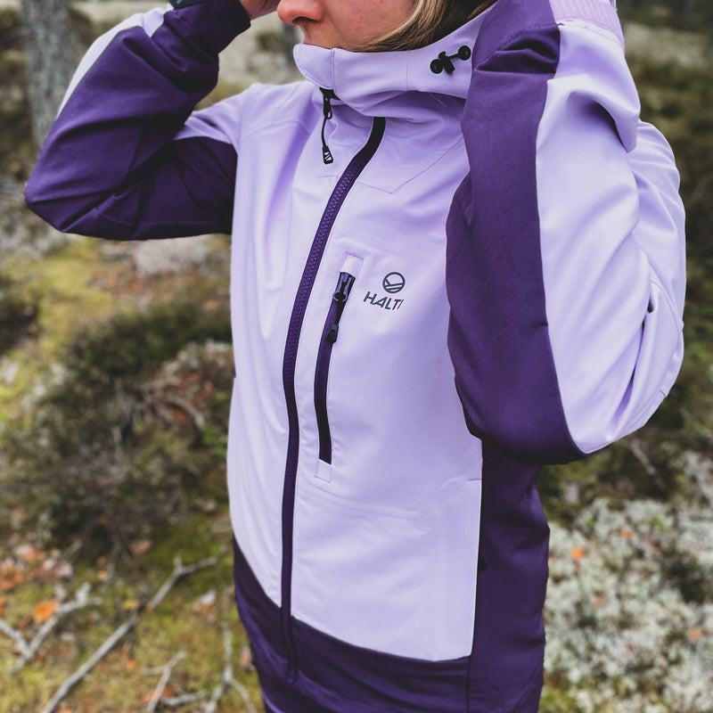 Person utomhus i en Halti Pallas III Dam Warm Hybrid Jacka, som justerar huvan med naturen suddig i bakgrunden.