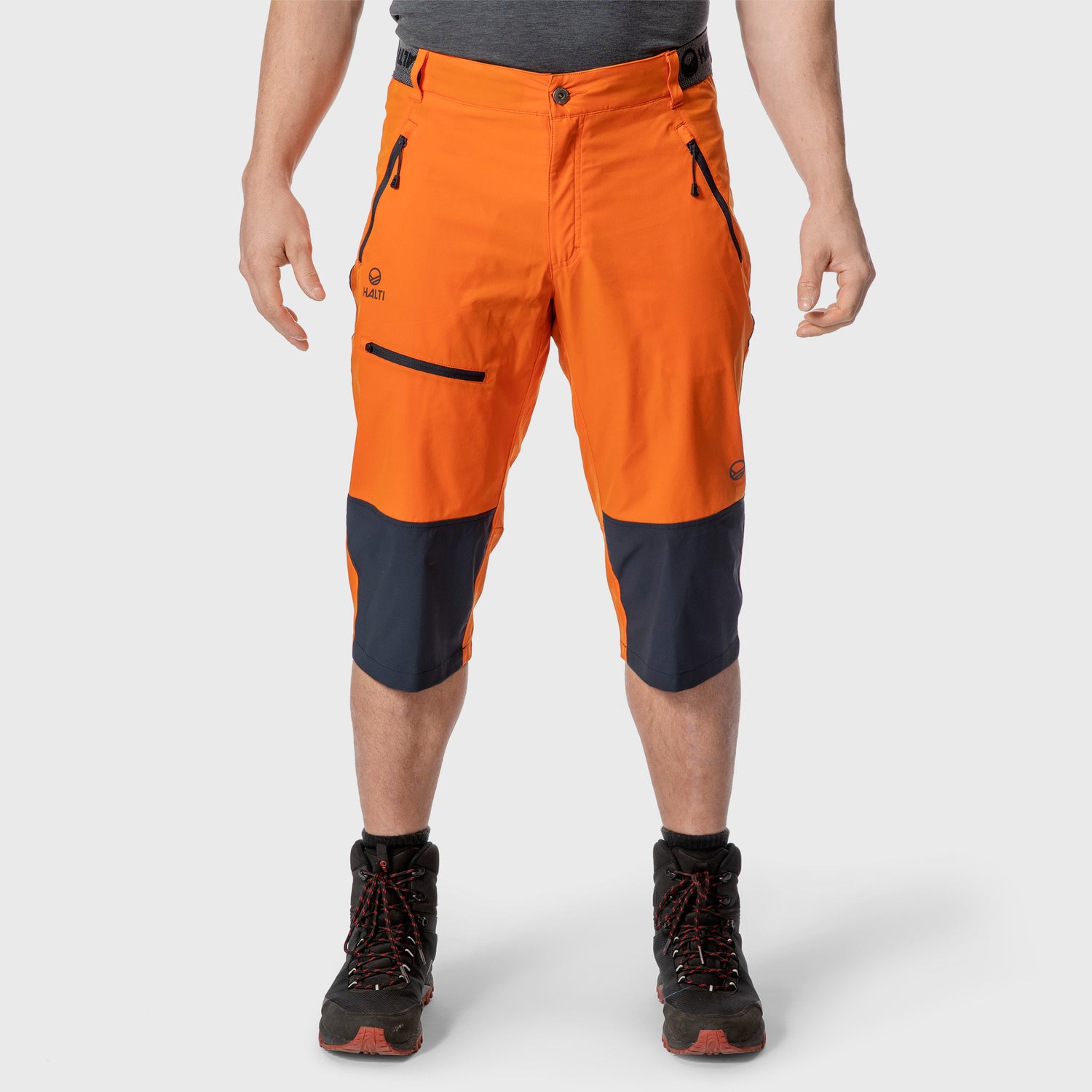 En person i ljusorange knälånga friluftsshorts med svarta partier och svarta vandringskängor står mot en enfärgad bakgrund.