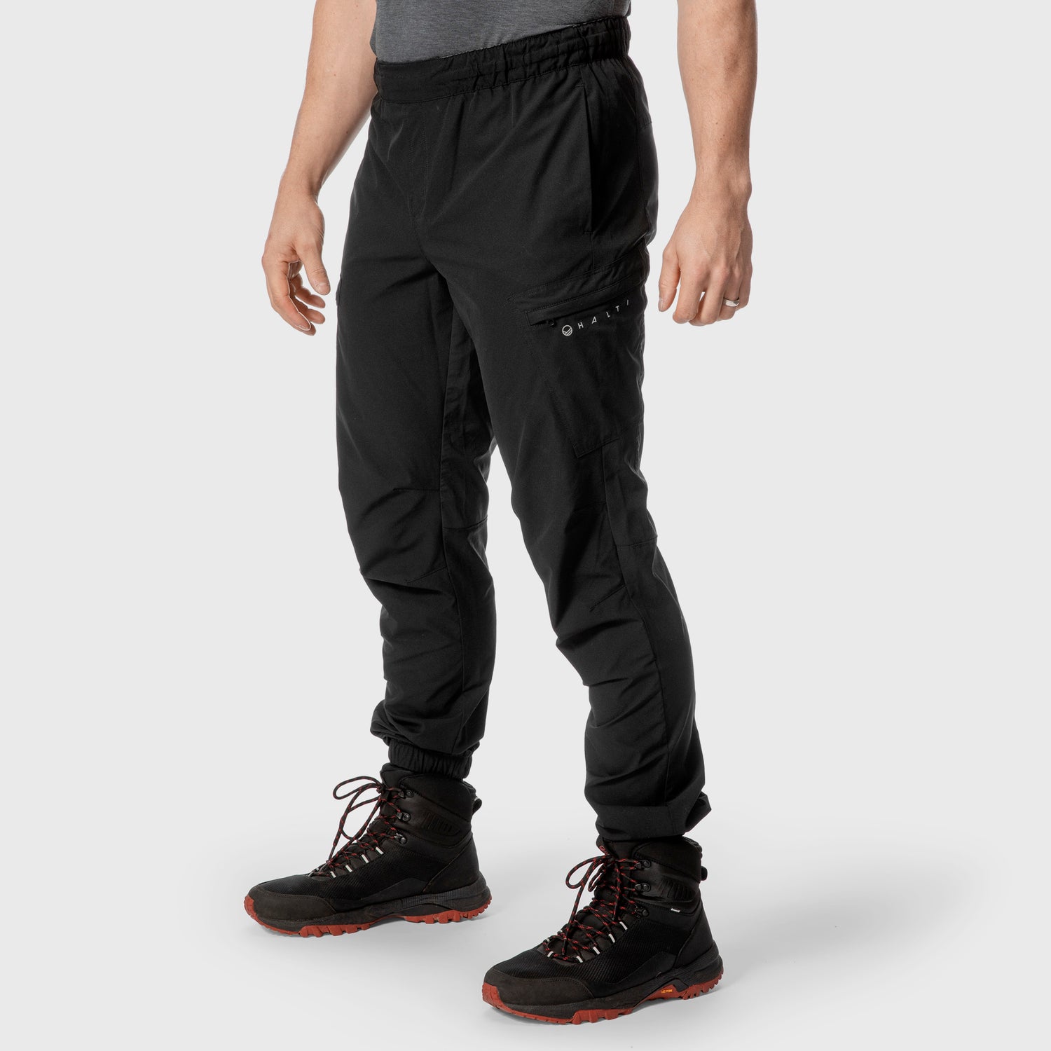 En person iförd svarta joggingbyxor, svarta vandringskängor med röda sulor och grå skjorta står mot en vit bakgrund.