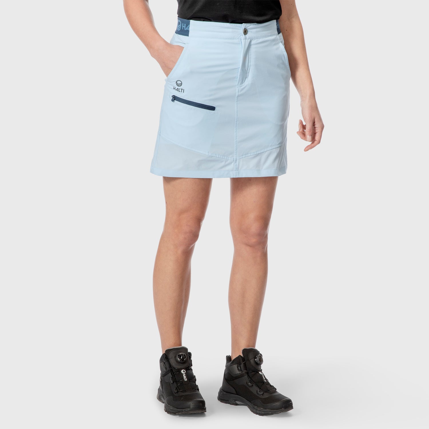 En person klädd i ljusblå kjol, svart skjorta och svarta vandringskängor står med ena handen i fickan.