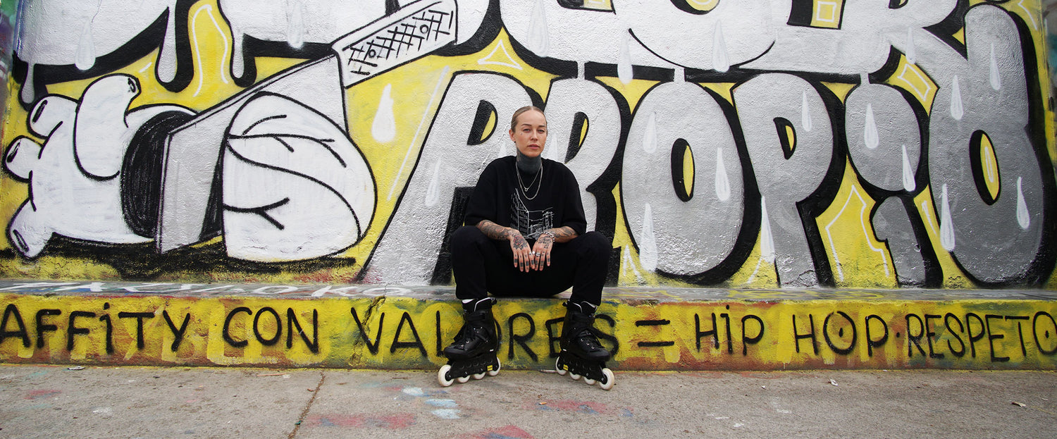 En mörkklädd person sitter på en avsats framför en färgsprakande graffitiklädd vägg med text och abstrakta mönster.