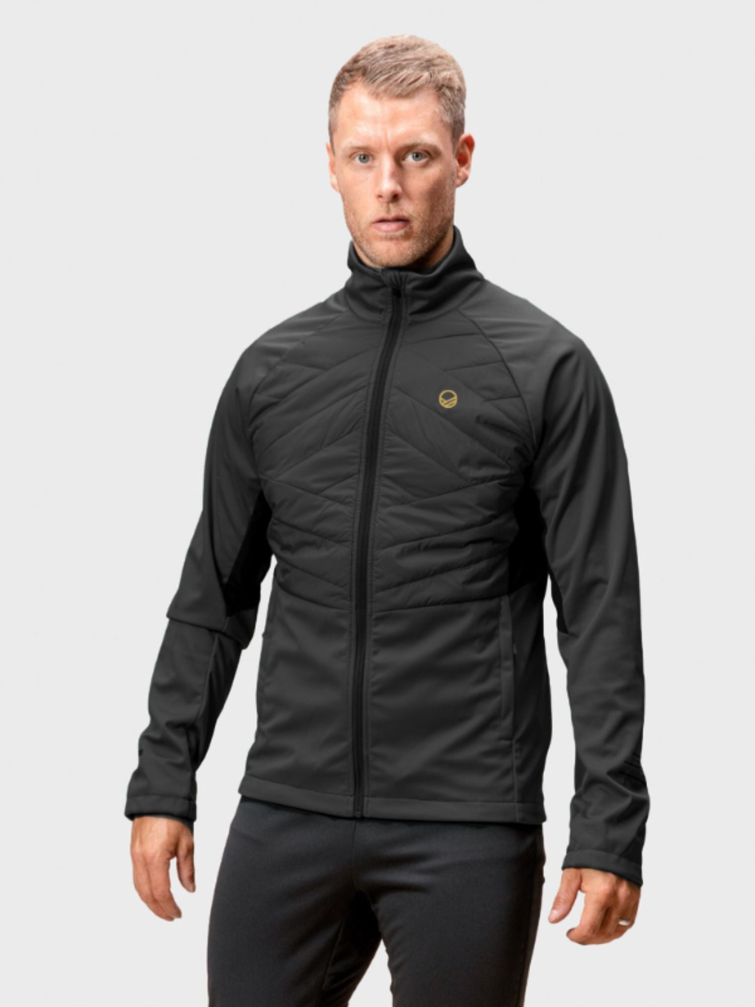 En man står i Halti Vinha Hybrid Längdskidjacka Herr och byxor, redo för vinterlöpning mot en ljusgrå bakgrund.