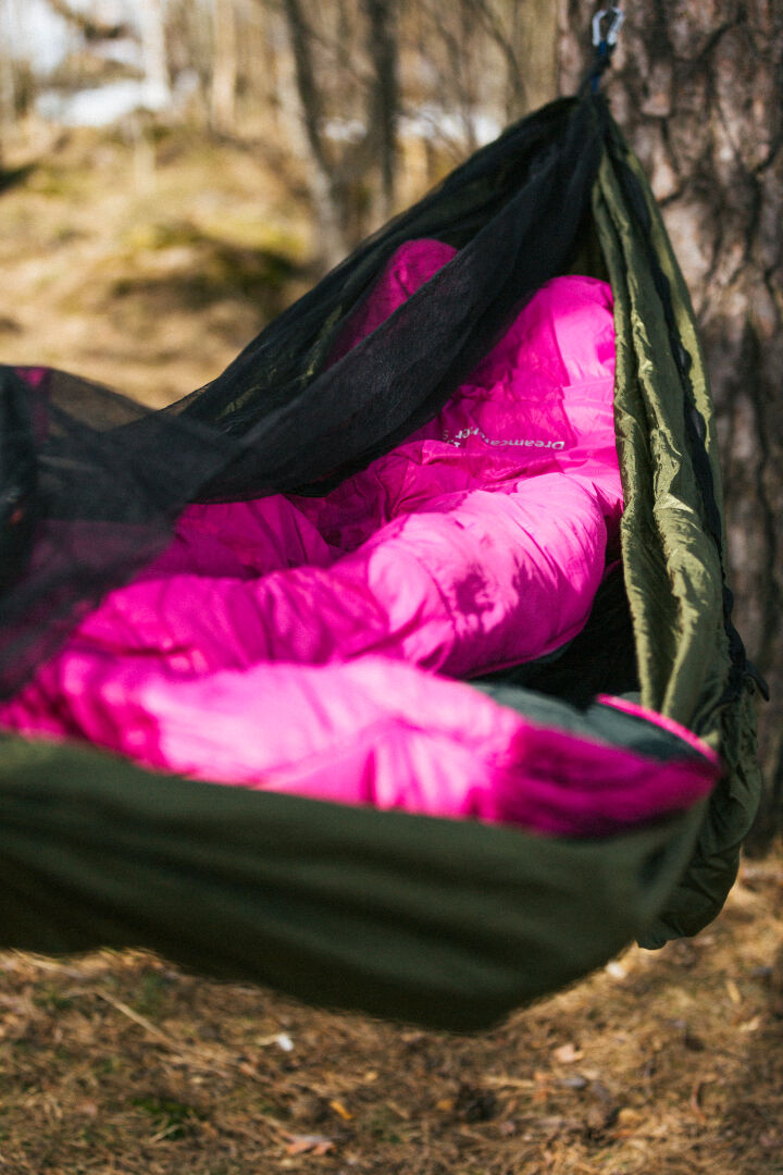 Någon i en knallrosa Halti Dreamcatcher 30 M sovsäck tillverkad av återvunnet material vilar i en grön hängmatta utanför.