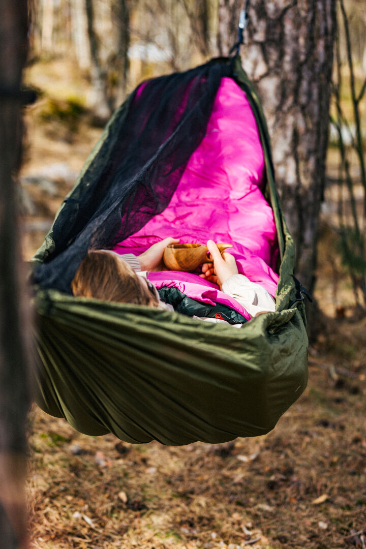 En person kopplar av i en grön hängmatta med en rosa filt och brun leksak, mysig i en Halti Dreamcatcher 30 M Sovsäck.