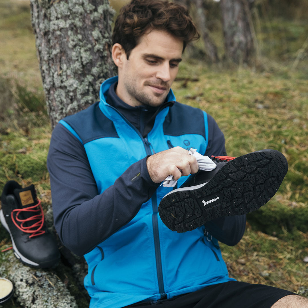 En man torkar bort lera från sin vandringssko; hans Halti Pallas Herr X-stretch Väst håller honom vatten- och vindtät.