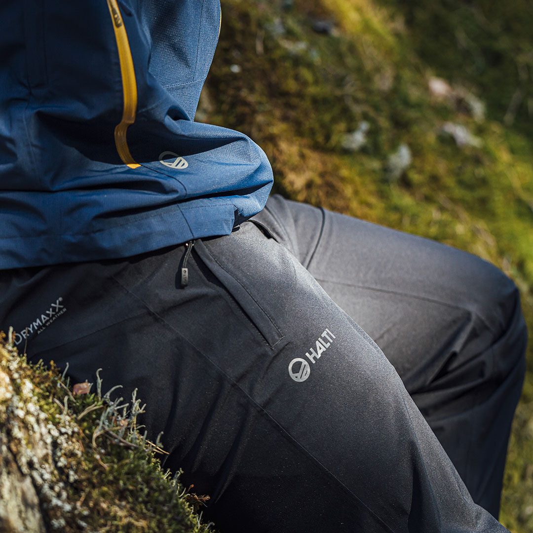 Person klädd i mörk Halti Vuoksi DX Skalbyxor Herr och blå jacka, sittande på mossig mark utomhus.