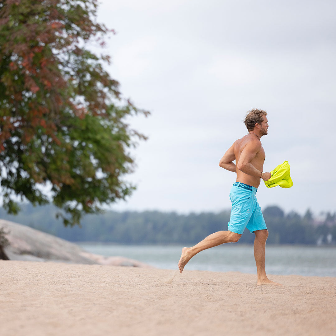 En man i Halti Pallas Herr X-stretch Lite Shorts springer barfota på en sandstrand med en gul skjorta i handen.