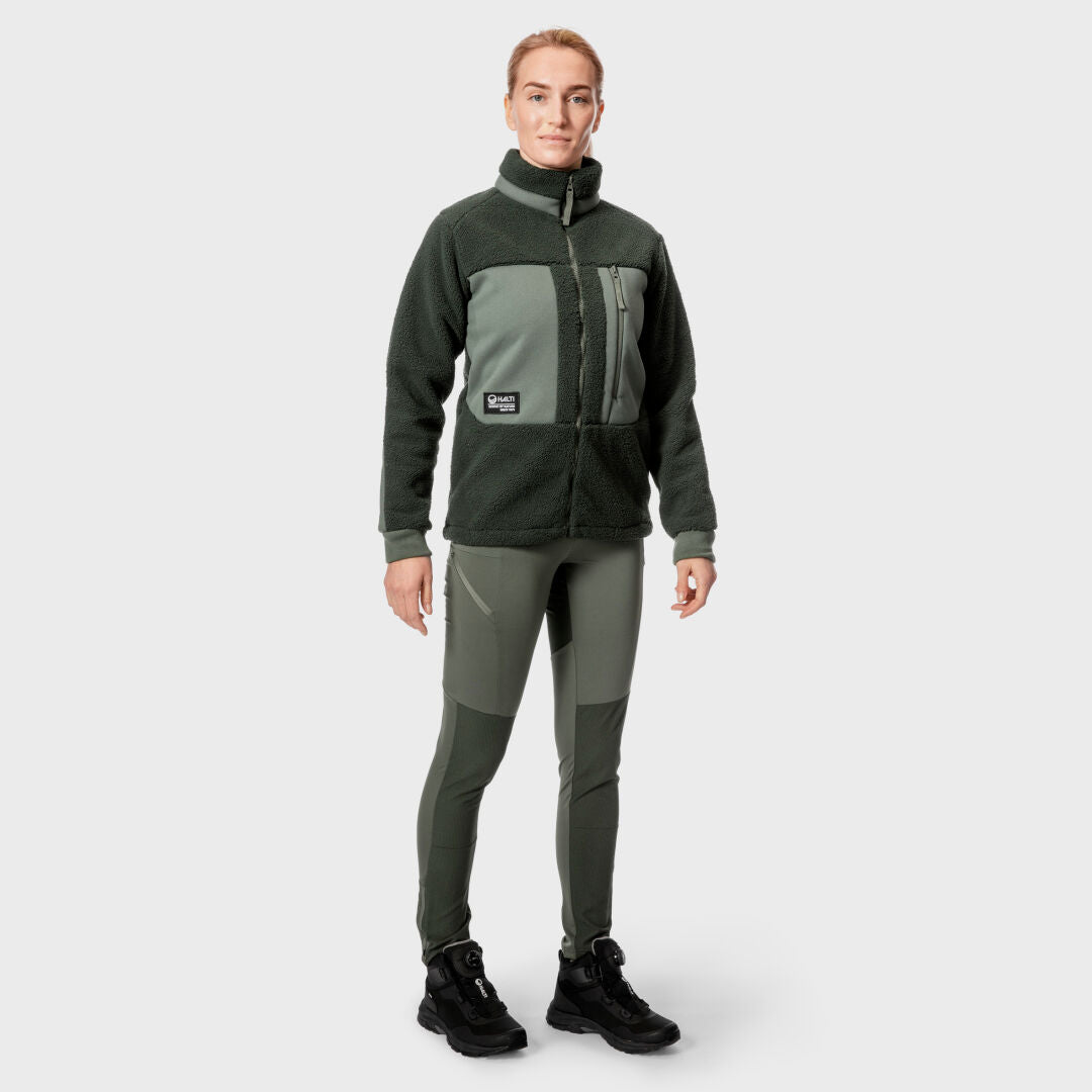 En person modellerar Halti Hiker Dam Polar Fleece med gröna byxor och svarta skor mot en enfärgad grå bakgrund.