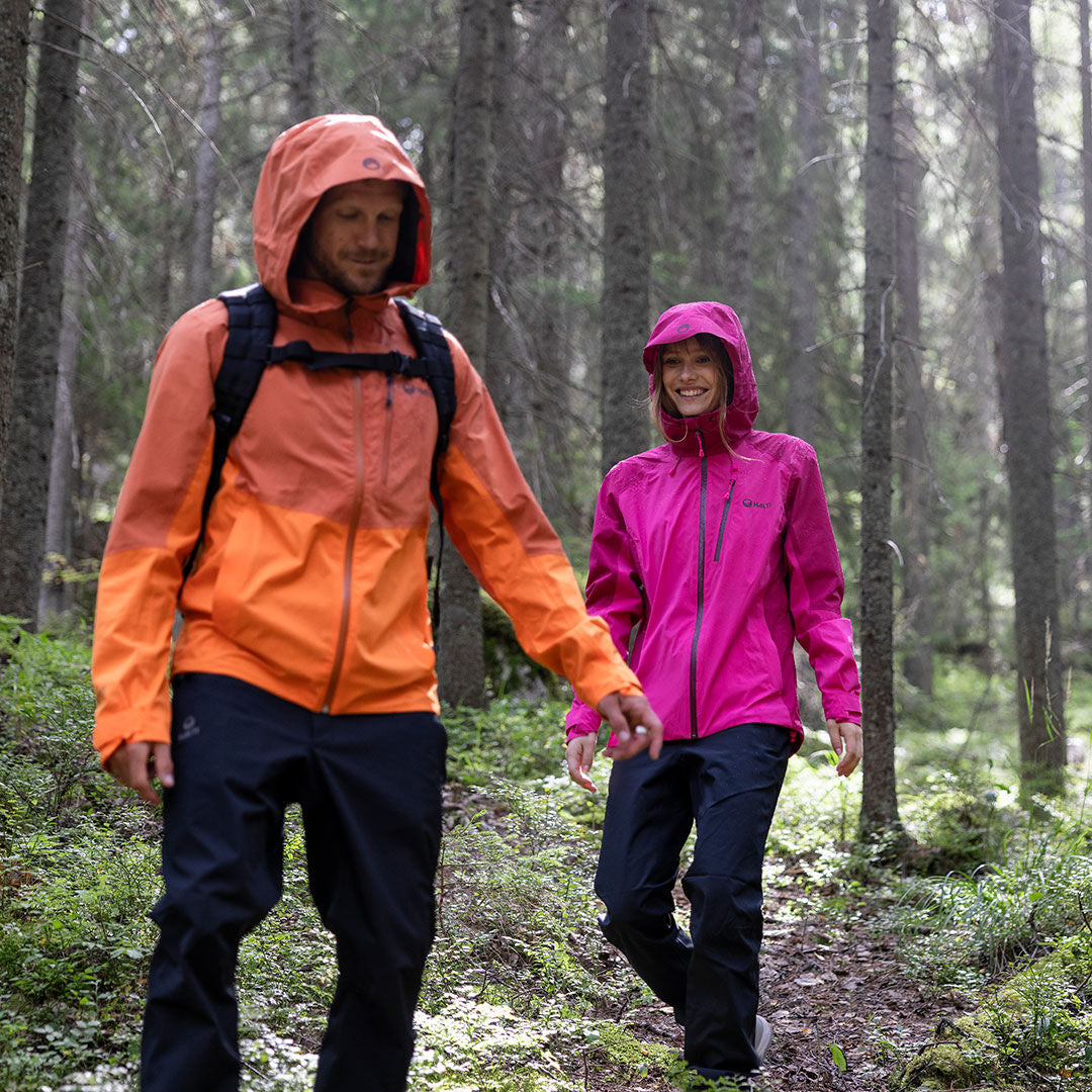 Två vandrare i Halti Vuoksi DX Skalbyxor vandrar på en grönskande stig - den ena i orange regnjacka, den andra i rosa.