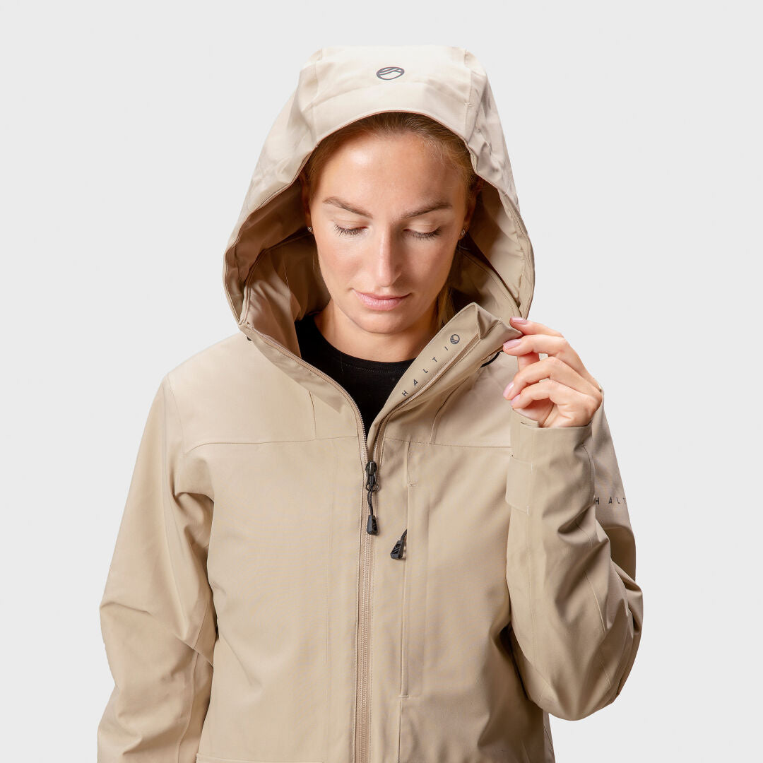 En person i en beige Halti Olhava DrymaxX Parka Dam står mot en ljus bakgrund, tittar ner och justerar dragkedjan.