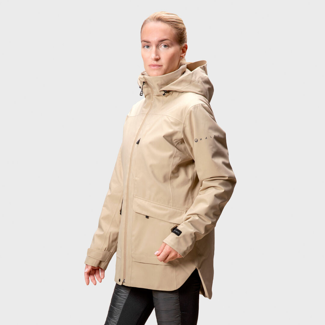 En person i en beige Halti Olhava DrymaxX Parka Dam och svarta byxor står mot en ljusgrå bakgrund.