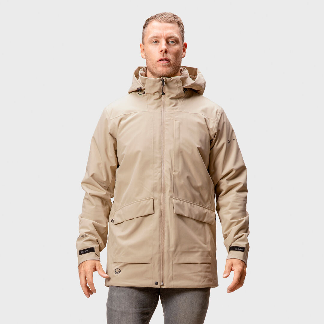 En man bär Halti Olhava DrymaxX Parka Herr i beige och grå byxor, stående mot en enfärgad ljus bakgrund.