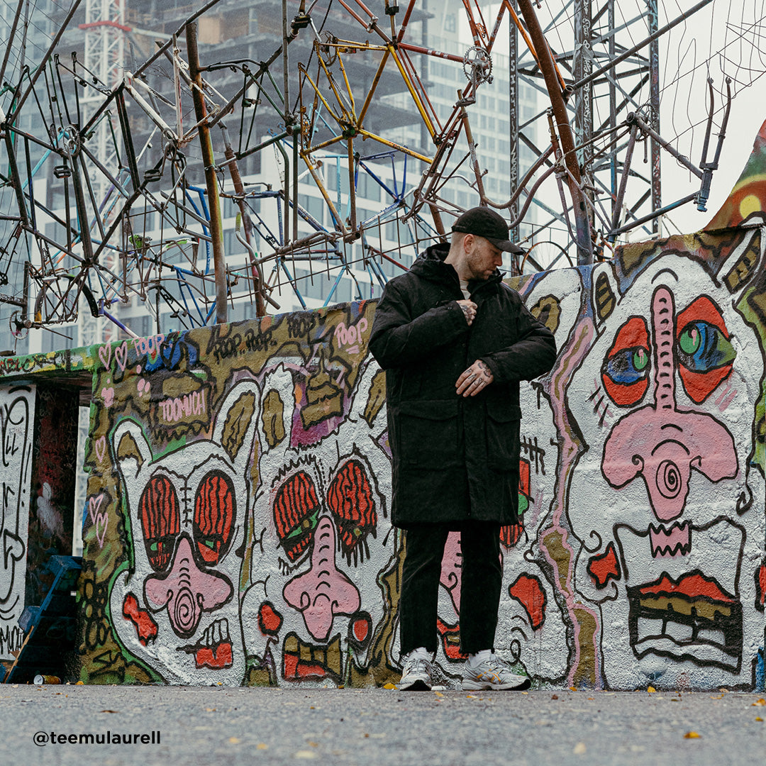 En person i en Halti Bergga Herr DrymaxX Vinterjacka står vid en graffitivägg med ansikten och abstrakt metall ovanför.
