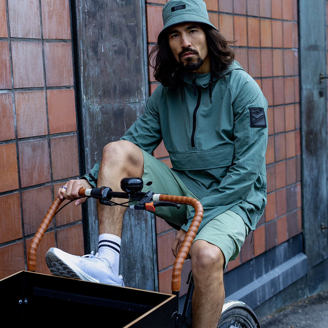 Man i Halti Blosis unisex-anorak och bucket hat sitter på en cykel med brunt styre vid en kaklad vägg.