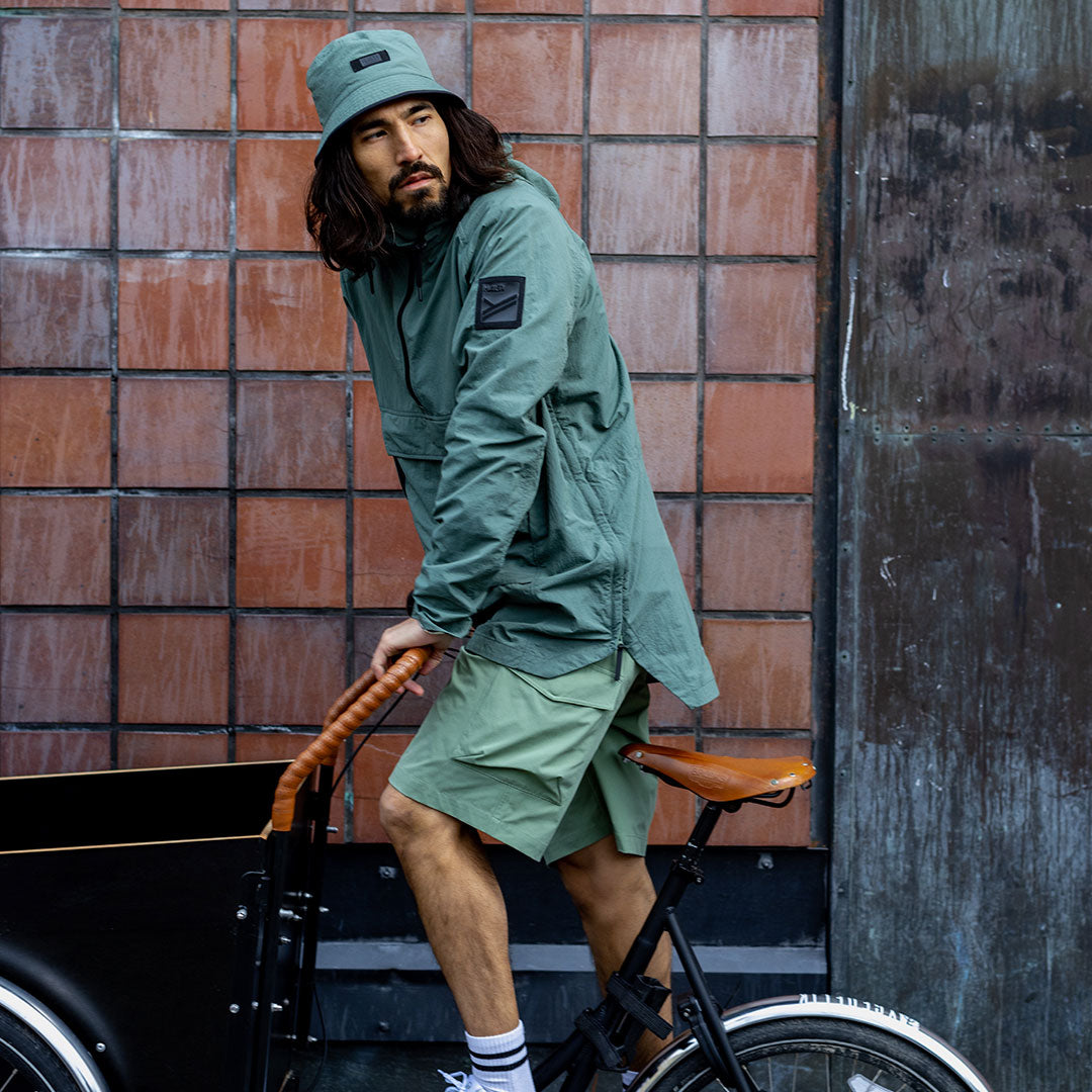 En person i Halti Blosis Unisex Anorak och bucket hat sitter på en cykel framför en kaklad vägg.