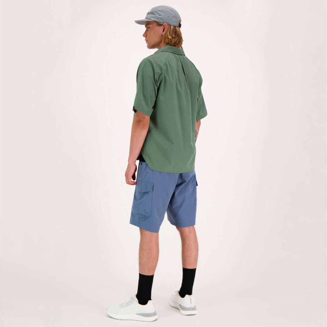 En person vetter bort och iklädd en Halti Baana Unisex Skjorta i gröna, blå seersucker-shorts och vita sneakers.