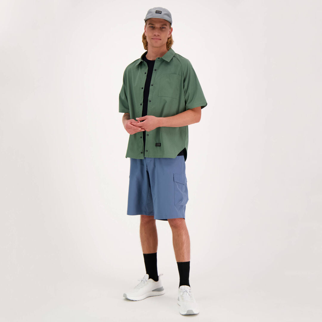 En person i Halti Baana Unisex Skjorta, blå shorts och vita sneakers står mot en enkel bakgrund.