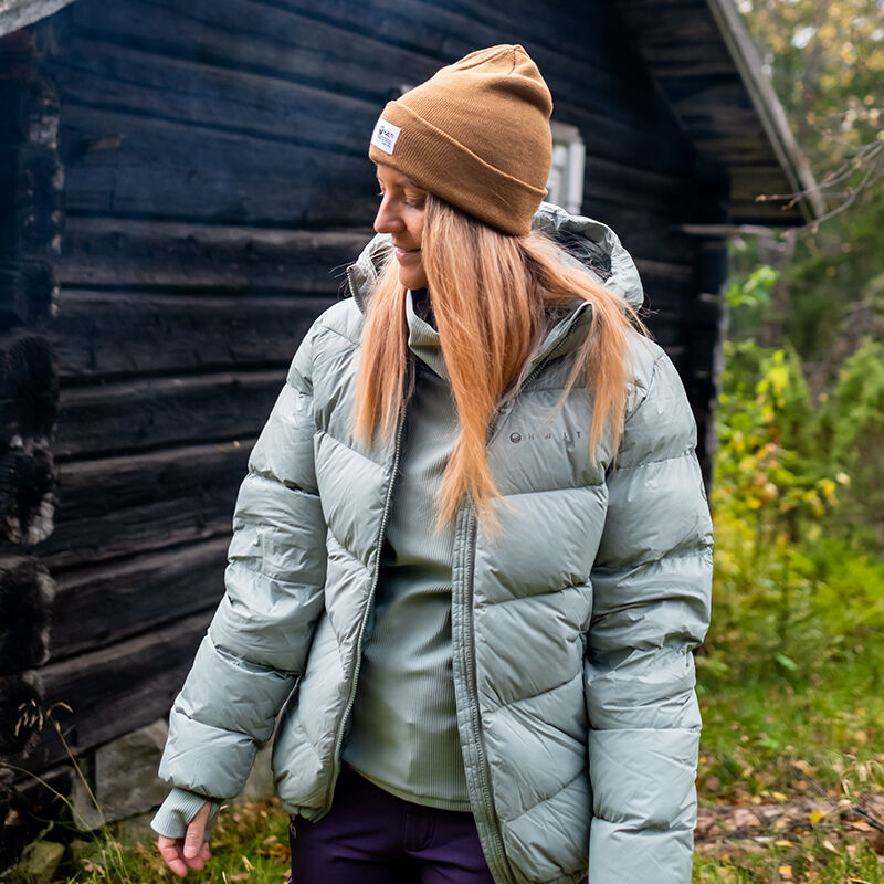 En person bär en Halti Versant Dunjacka Dam i ljusgrått och en brun mössa utanför vid en trästuga med grönska.