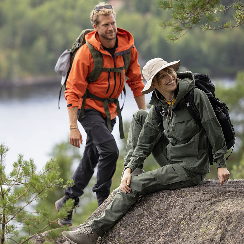 Två personer i Halti Hiker Dam Ventilated Byxor vandrar vid en sjö; den ena sitter på en klippa, den andra står bakom.