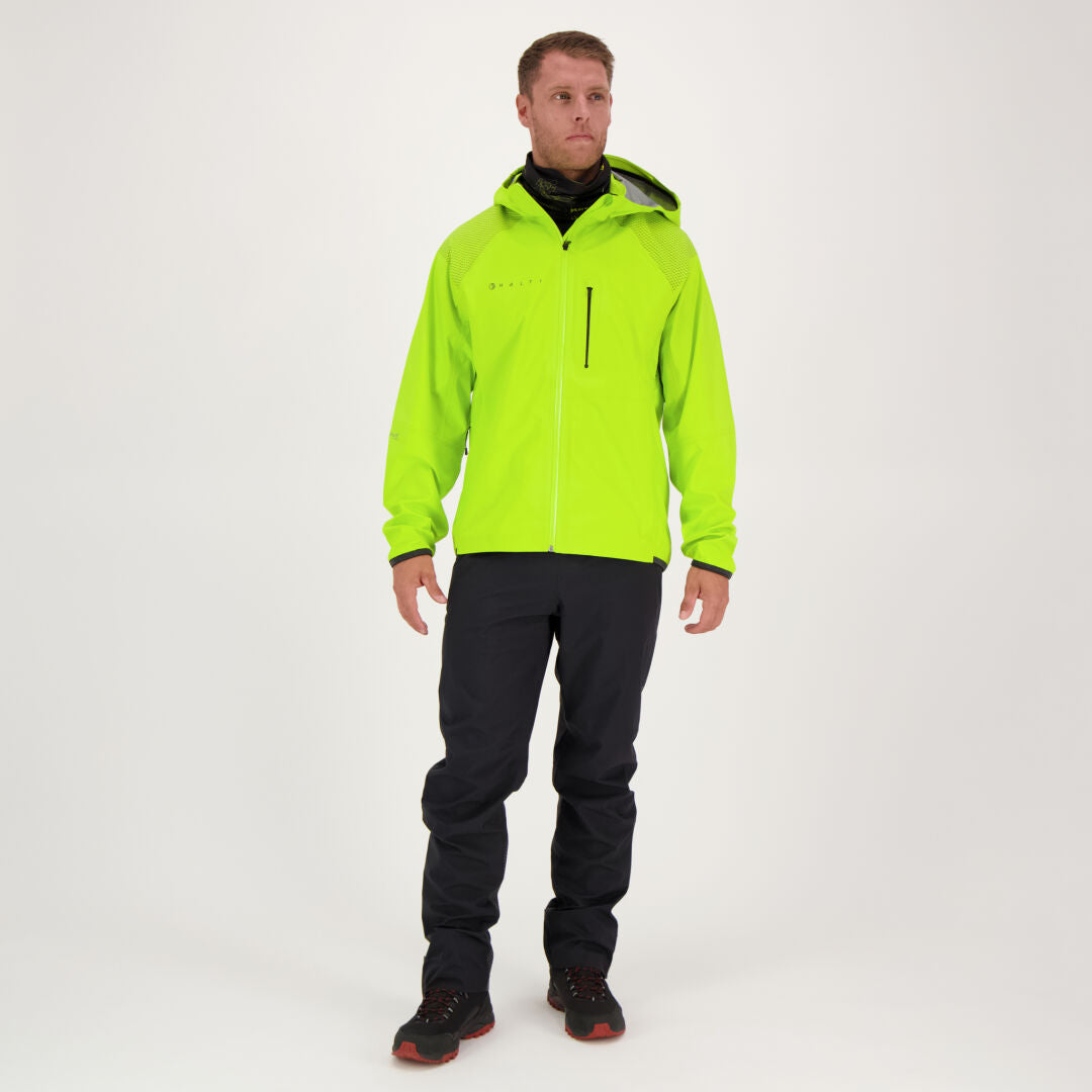 En man står i en studio iklädd en klargrön luvjacka, svarta Halti Liike Herr 3L DrymaxX Sport Byxor och svarta skor.