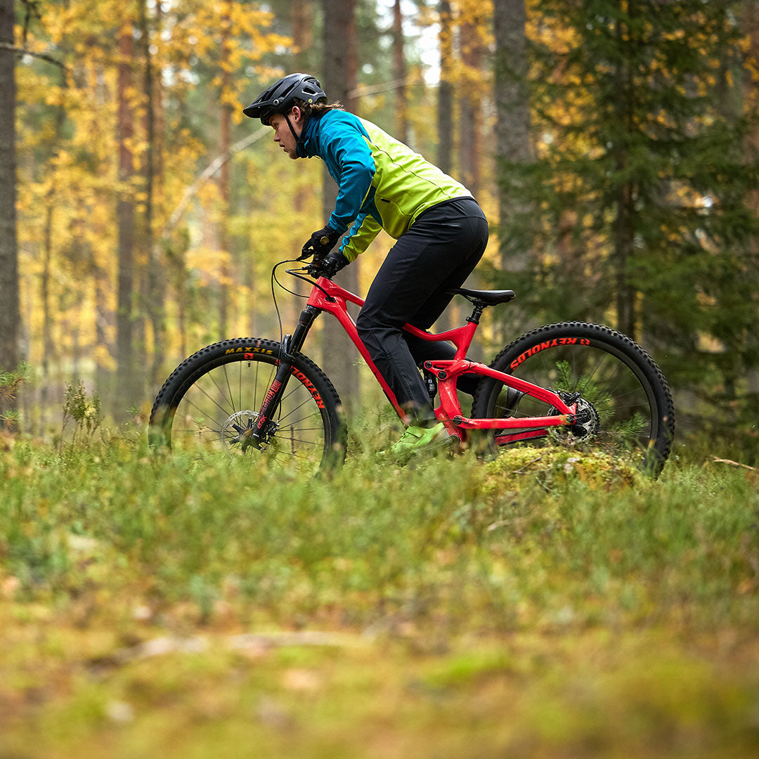 En person i en Halti Suunta Herr Hybrid Jacka cyklar på en röd mountainbike på en skogsstig med höstträd.